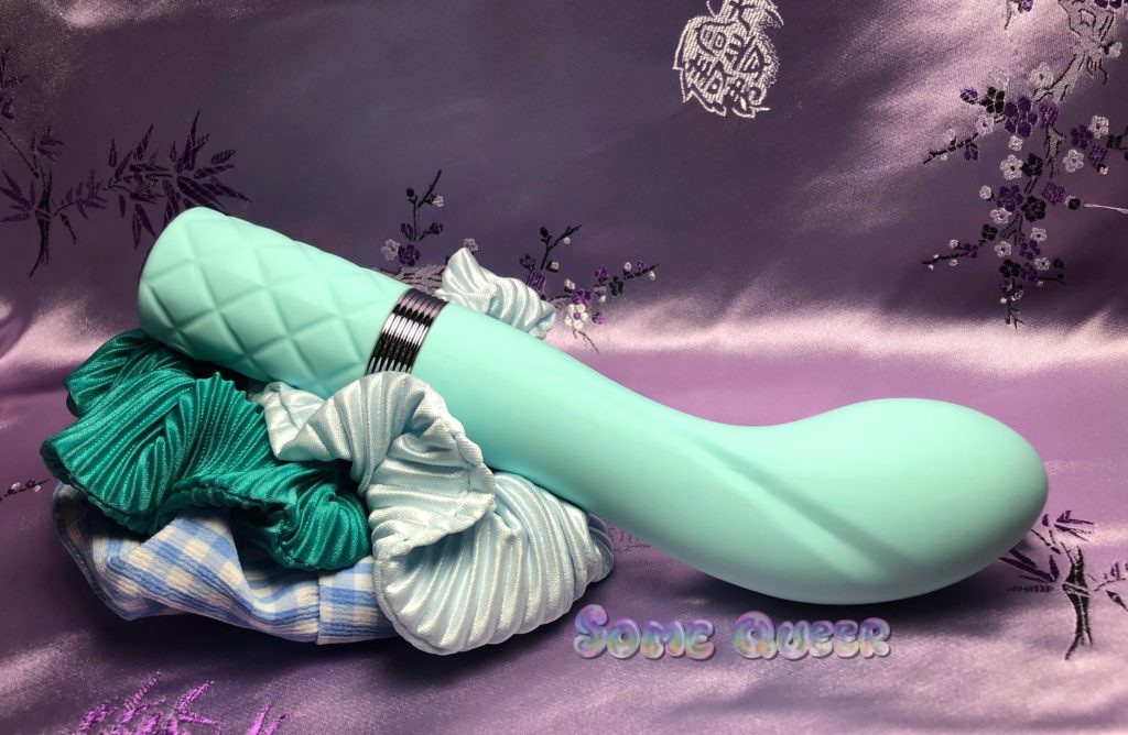 Sassy by Pillow Talk, in the teal color, propped on top of various shades of blue fabric. The background is purple with small floral patterns.