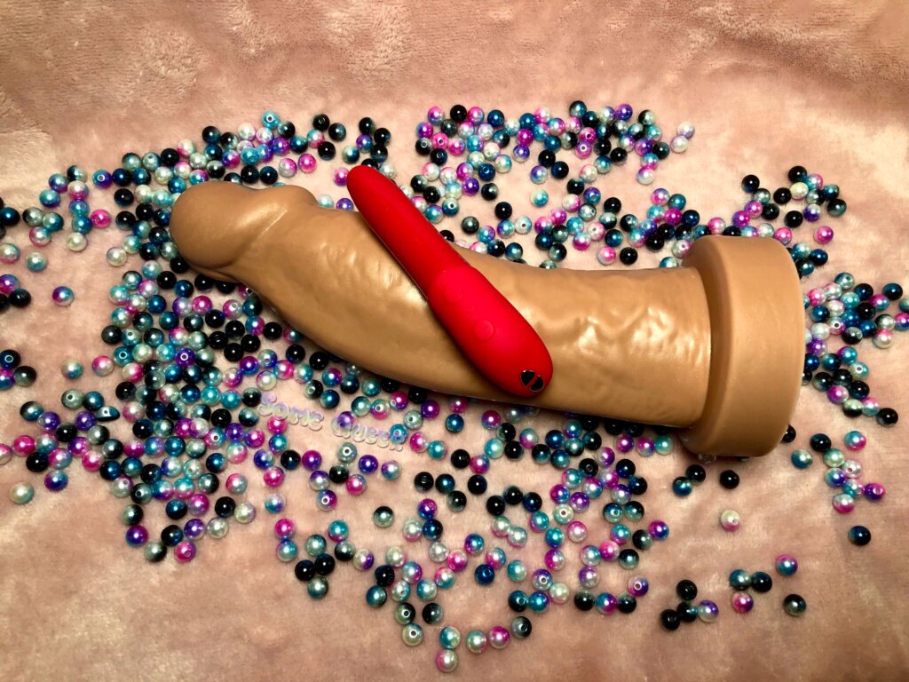 Dee's Big Daddies Diedrich laying on top of multicolored beads, and a pink background. There is a red We-Vibe Tango X vibrator laying on top of it.