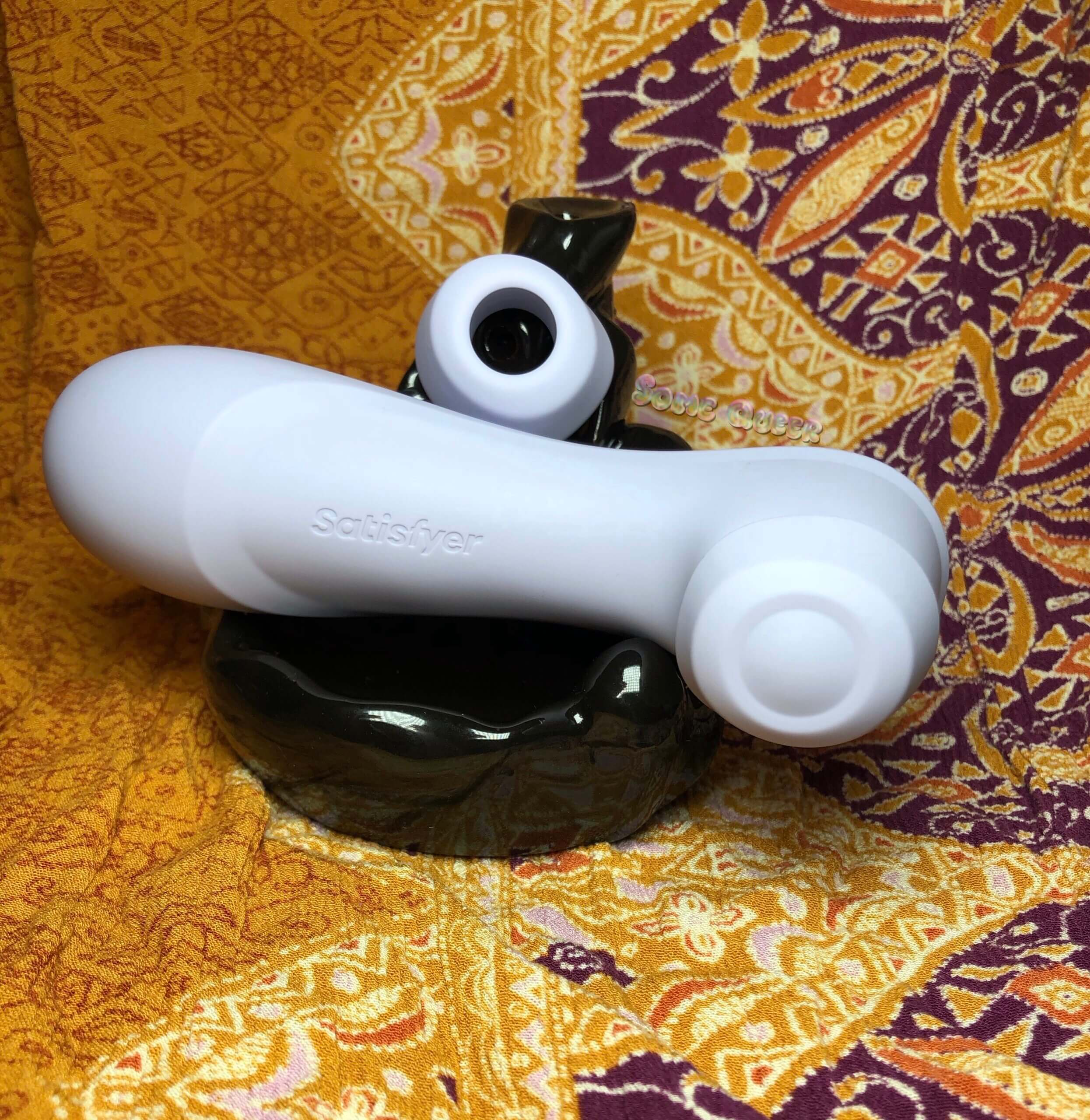 Satisfyer Pro 2 Gen 3 in lilac with liquid air cap attached, and classic air pulse cap resting on top. The toy is sitting in a shiny black decor piece.