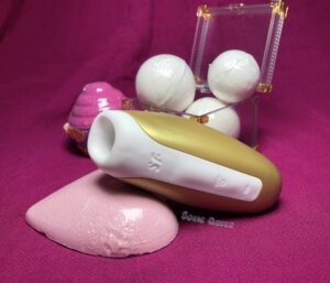 Satisfyer Love Breeze in yellow surrounded by bath bombs, all set on a magenta background.