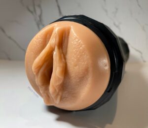 Front view of the Cupsland Twirl 1.0 It is a skin colored T.P.E vulva inside a black plastic case. The product is set on a white marble background. 