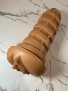 T.P.E sleeve of the Cupsland Twirl 1.0 laying on a white marble background. The sleeve is a tan flesh color, and has a swirl design on the outside, with a vulva shaped front. 