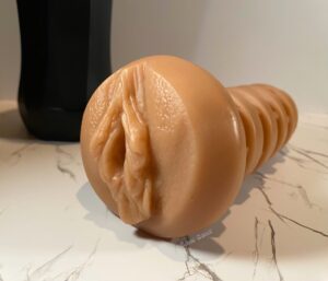 Vulva shaped sleeve from the Twirl 1.0 laying on a white marble background. The black case of the stroker is out of focus in the background. 