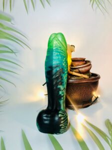 Underside of the Fascinos dildo from Uberrime in front of a decorative statue of candles and vases. The dildo is shades of blue, green, and black