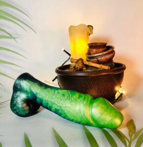 Fascinos dildo from Uberrime in front of a decorative statue of candles and vases. The dildo is shades of blue, green, and black.