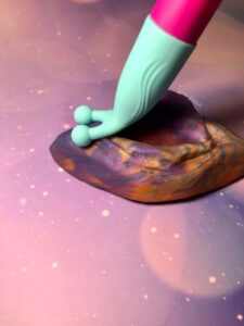 The maci vibelite maci cupping the clitoris of a silicone vulva squish. The vulva squish is blue, orange, and purple marbled together.