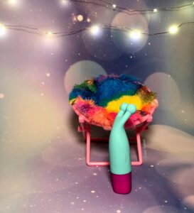 Side profile of the Maia Vibelite Maci. The teal and magenta vibrator is turned sideways, standing in front of a fuzzy, miniature tie-dye chair.