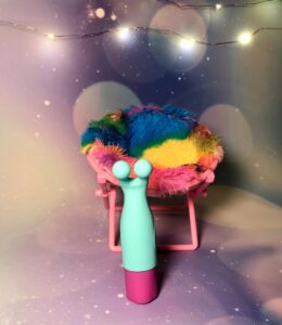 The Maia vibelite maci. A small teal and magenta vibrator. It has two rounded prongs for the end. It's standing in front of a miniature, fuzzy tie-dye chair.