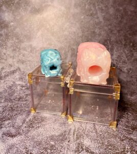 The Falkor stroker and Falkor fidget squish sitting side by side on clear boxes with gold corners. The squish version is blue and black, and the full size is rose quartz colored. 