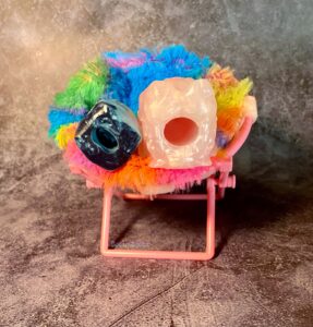 The Falkor stroker and the Falkor squish sitting together in a miniature, colorful, fuzzy chair. The full size Falkor is forward, and the squish Falkor is backwards, so the openings can be compared. 