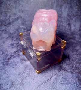 Falkor Micro Stroker from Squidly Co in rose quartz coloration. The Falkor stroker has a dragon maw appearance. The silicone stroker is sitting on top of a clear box with gold corners. 