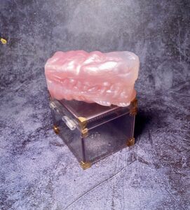 Falkor Micro Stroker from Squidly Co in rose quartz coloration. The Falkor stroker has a dragon maw appearance. The silicone stroker is sitting on top of a clear box with gold corners. 