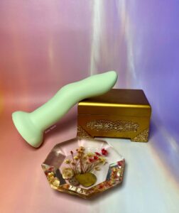 The Crassie dual density strap on dildo laying on top of a small, gold box. There is a geometric, clear resin flower pour in front of them. The entire background is sunset colored.