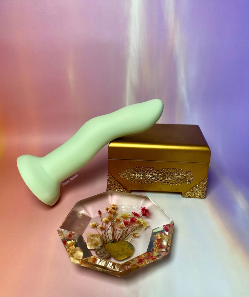 Crassie dual density dildo in green laying on a small, gold jewelry box. There is a clear resin Knick knack with flowers inside laying in front of it all.