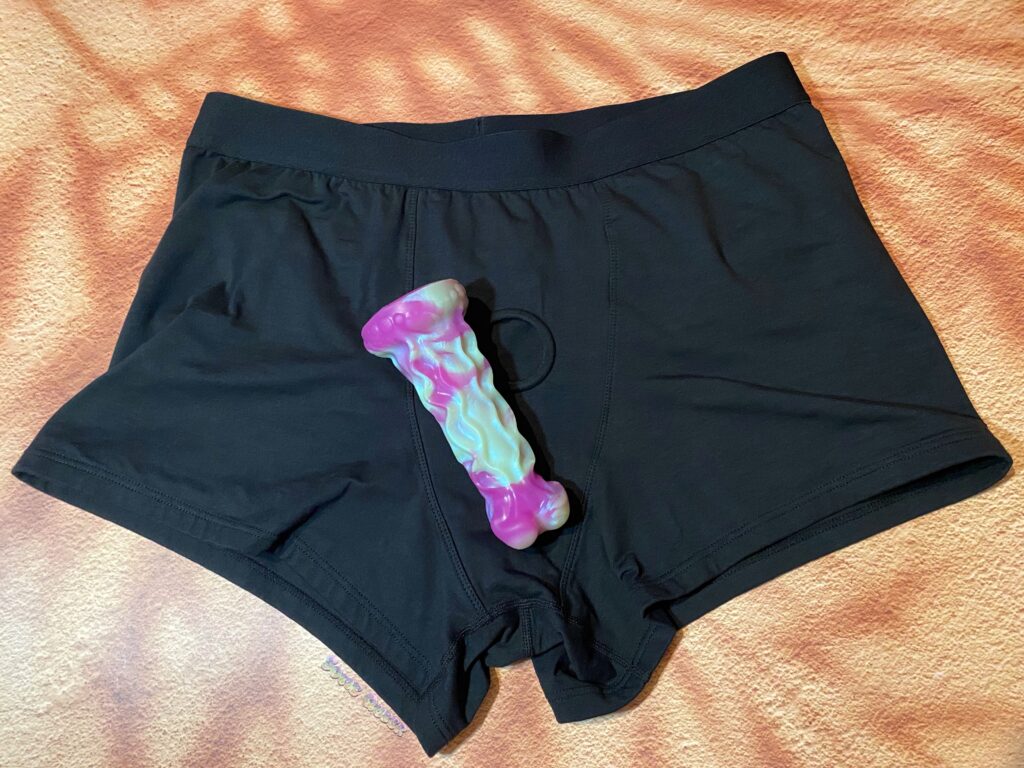 Black SnugHer boxer harness laid flat with a purple and green textured dildo laying on top