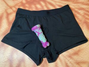 Front shot of the SnugHer boxer harness with the same green and purple textured dildo laying on them. 