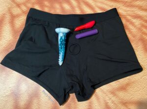 Front shot of the SnugHer boxer harness with a tentacle dildo and two bullet vibes, one red, one purple, laying on top. 