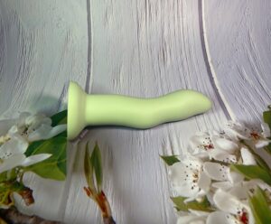 The Crassie Dual density strap on dildo in green laying on a white wooden background with flower blossoms.