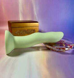 The Crassie dual density strap on dildo laying on top of a geometric, clear resin flower Knick knack, a small gold jewelry box behind them. The background is sunset colored