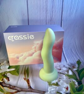 The Crassie Box with the green dual density strap on dildo in front of it. The box is a pink and purple sunset sky full of clouds.