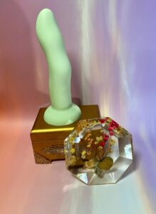 The green Crassie dual density strap on dildo standing on top of a small, golden jewelry box, with a clear resin and flower Knick knack propped against the box.