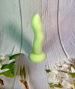 The green colored, Crassie dual density strap on dildo standing upright on a white, wooden colored background, with flower blossoms on it.