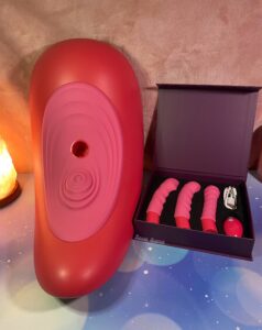 The Rider Desire saddle and box of attachments beside it again. The box is a purple box with a black foam cutout to hold everything. The Rider Desire is described as "rose red" by the company, which is more of a pink color.