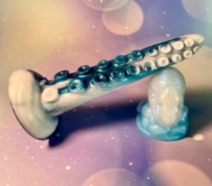 Thalia Tentacle propped on top of Squidly Co's nautilus squish. Thalia is a tentacle dildo in white and shimmery blue coloration. The nautilus squish is white and light colored blue. 